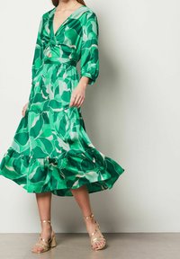 Kvinna iklädd en grön blommig midiklänning med trekvartsärmar och guldstrappy blockklacksandaler, stående mot en enfärgad vägg.