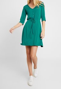 Robe verte à manches courtes en tissu doux, avec un décolleté en V, un devant à boutons, une taille à cordon et un ourlet évasé. Portée avec des baskets blanches.