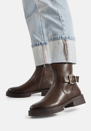 Ciemnobrązowe skórzane botki z klamrą, noszone z jasnoniebieskimi podwiniętymi dżinsami na jednolitym tle.