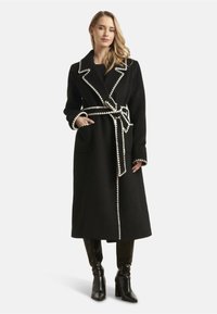 Long black wool coat with white contrasting stitching, notched lapels, side pockets, and a fabric belt, paired with black shiny heeled boots.