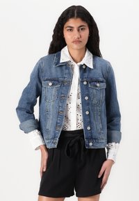 La chaqueta de mezclilla presenta un corte clásico en azul medio, con cierres de botones y bolsillos en el pecho. Combinada con una camisa blanca y pantalones cortos negros.
