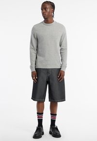 Pull gris à manches longues, shorts en denim noir avec des coutures visibles, chaussettes noires et rouges avec du texte, et chaussures noires à lacets.