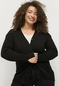 Zwarte cardigan van een zachte gebreide stof met een V-hals, lange mouwen en knoopsluiting. Heeft ribgebreide manchetten en zoom.
