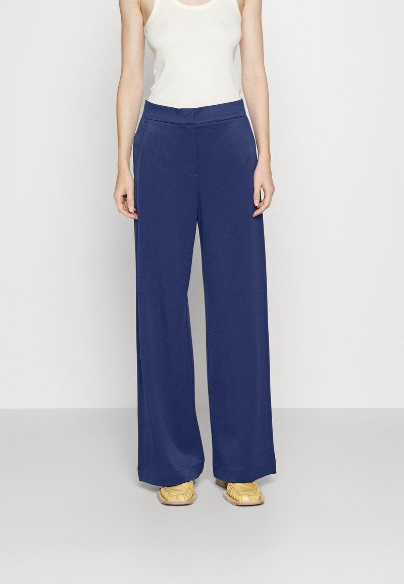 Femme portant un pantalon large taille haute bleu marine associé à un haut sans manches blanc et des mocassins jaunes, debout devant un fond uni.