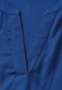 Primo piano di un collo di un indumento blu con apertura a forma di V e dettagli di cucitura visibili.