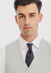 Szary kamizelka w wzory, noszona na białej koszuli, z ciemnoniebieskim krawatem we wzór geometryczny. Gładka faktura i dopasowany krój.