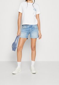 Vit croppad t-shirt med en teal logotyp, ljusblå denimshorts, vita sneakers och en blå mönstrad handväska.