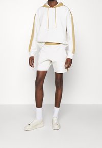 Vit hoodie med guld detaljer och dragsnören, i kombination med matchande vita shorts. Bärs med vita strumpor och krämfärgade sneakers.