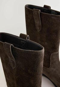 Bottines en daim marron avec une texture lisse, des têtes arrondies et des languettes sur les côtés pour un enfilage facile. La doublure intérieure est noire.