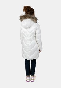 Giacca a vento bianca imbottita con trapuntatura a diamante e cappuccio bordato di pelliccia, abbinata a jeans scuri e sneakers colorate. Vista da dietro.