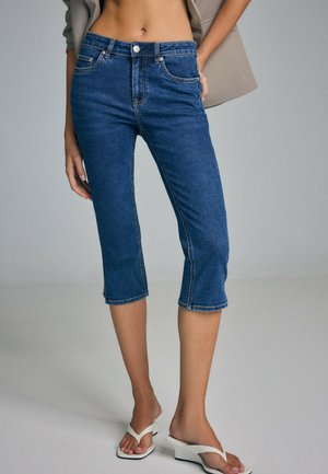 Femme vêtue d'un jean court en denim bleu moyen, d'un blazer beige et de sandales blanches à talons, debout avec une main dans la poche.