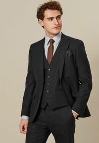 Dunkelgrauer Dreiteiler bestehend aus einem taillierten Blazer, einer Weste mit Knöpfen und einer Hose. Begleitet von einem hellblauen Hemd und einer braunen Krawatte.