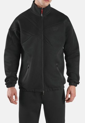 Homme portant une veste noire zippée avec des motifs cousus subtils et des poches latérales zippées, associée à un pantalon noir assorti.