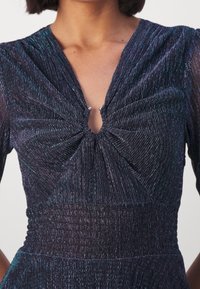 Femme portant un haut plissé métallique foncé avec un détail noué froncé au col en V et une taille froncée.