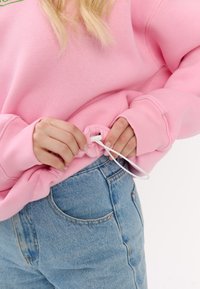 Felpa rosa chiaro con coulisse all'orlo, abbinata a jeans in denim blu. La felpa presenta polsini a costine e una texture liscia.