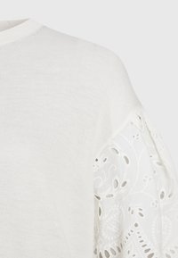 Biała dzianinowa bluzka z okrągłym dekoltem i rękawami ozdobionymi delikatnymi, białymi haftowanymi wzorami w kwiaty.