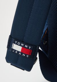 Sangle en tissu bleu marine avec une surface texturée, dotée d'une patch rectangulaire affichant le logo "TOMMY JEANS" en lettres blanches et rouges.