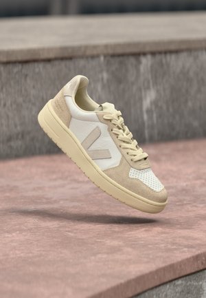 Beige en witte lage sneaker met suède en leren panelen, geperforeerde neus en een distinctief "V"-logo aan de zijkant.