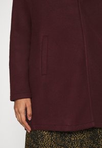 Bordeauxkleurige mantel van een wollen mix met een minimalistische structuur, voorzien van een zijzak en een zachte textuur. De onderrand is netjes afgewerkt.