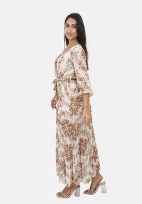Robe maxi en crème avec motif floral marron, manches longues, décolleté en V, taille nouée, texture fluide, portée avec des talons transparents.