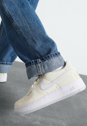 White leather Nike sneakers with a textured finish, featuring a grey swoosh logo, white rubber sole, and perforated toe box, paired with blue jeans.