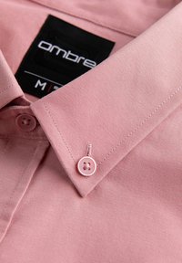 Gros plan sur le col d'une chemise rose à boutons avec un bouton assorti et une étiquette de marque noire indiquant « ombre » et taille M.
