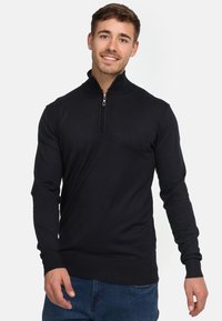 Suéter negro de punto con cuello de media cremallera, tejido texturizado, mangas largas y puños acanalados. Lleva puesto sobre unos vaqueros de mezclilla azul.