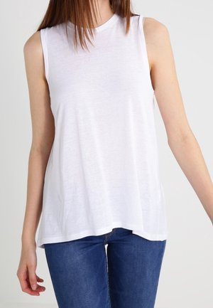 Kobieta ubrana w bezrękawnik biały top w połączeniu z niebieskimi dżinsami na jednolitym jasnym tle.