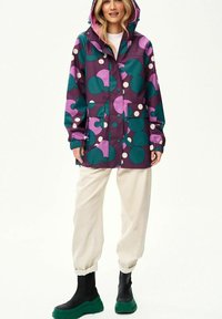 Kobieta ubrana w kaptur deszczowy z wzorem w kółka w kolorach fioletowym, różowym, turkusowym i białym, białe spodnie i czarne buty z zielonymi podeszwami, stojąca.