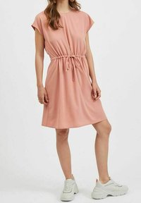 Lososově růžové šaty s krátkými rukávy a elastickým stahovacím páskem v pase; měkká, hladká látka; délka ke kolenům; doplněné bílými teniskami.