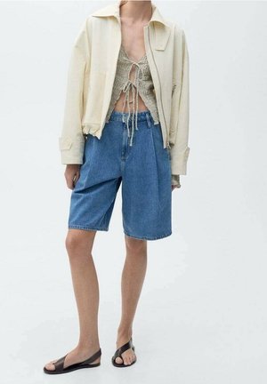 Persoon draagt een crème korte jas, een gebreide crop top met strikjes aan de voorkant, losse blauwe denim shorts en zwarte platte sandalen, staand tegen een witte achtergrond.