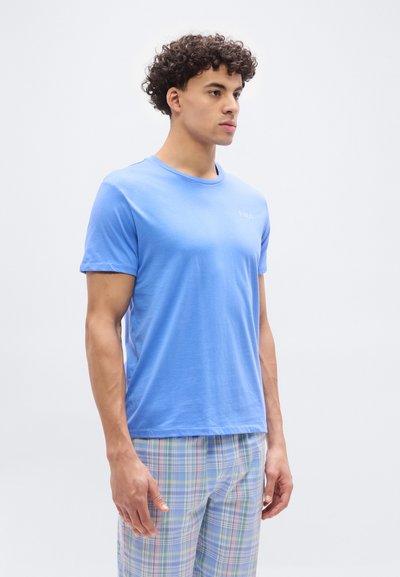 Νεαρός άνδρας με σγουρά μαλλιά, που φορά ένα ανοιχτό γαλάζιο μπλουζάκι Polo και παστέλ καρό  παντελόνι, στέκεται μπροστά σε λευκό φόντο.