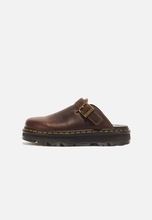 Zapato de cuero marrón tipo slip-on con suela negra gruesa, costuras amarillas y una correa con hebilla dorada ajustable en el lateral.