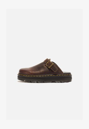 Zapato de cuero marrón tipo slip-on con suela negra gruesa, costuras amarillas y una correa con hebilla dorada ajustable en el lateral.