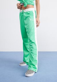 Green athletic pants made of smooth fabric with white side stripes and a drawstring waist, paired with black and white checkered shoes.