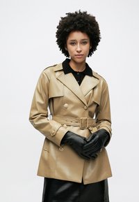 Μπεζ παλτό τύπου τρανς από ψεύτικο δέρμα με διπλή σειρά κουμπιών, μεγάλο γιακά, ζωνάκι στη μέση και μαύρα γάντια. Ομαλή υφή με επιχρυσωμένες λεπτομέρειες.