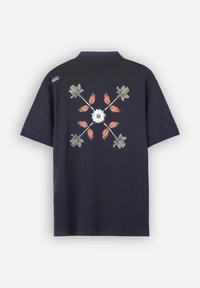 Polo-shirt à manches courtes bleu marine avec un motif floral central comprenant une marguerite blanche et des feuilles colorées. Comprend un petit logo sur le col.