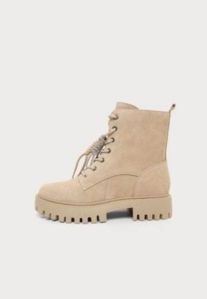 Stivaletto beige in suede con lacci alla caviglia, suola in gomma spessa e linguetta di tiraggio su sfondo chiaro uniforme.