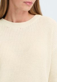 Maglione in maglia color crema chiaro con un motivo testurizzato, scollatura rotonda e polsini a costine. Il tessuto sembra morbido e traspirante.