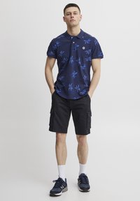 Marineblå poloshirt med blå blomsterdesign, krage og logodetaljer; kombinert med svarte cargo shorts og marineblå treningssko. Hvite sokker synlige.