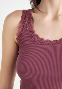 Top a maniche senza maniche color malva con dettagli in pizzo floreale lungo il colletto e le spalline, caratterizzato da una vestibilità aderente e una texture morbida.