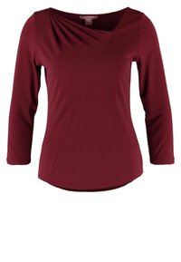 Burgundy long-sleeve top with a draped neckline, asymmetrical front seam, and a rounded hem. Made from smooth, stretchy fabric.
