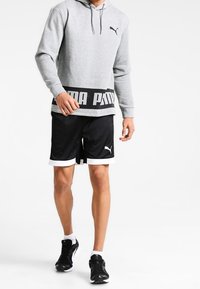 Grå hoodie med svart logoband, matchad med svarta shorts med vit kant. Modellen bär svarta träningsskor.