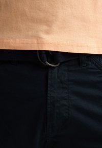 Maglietta in cotone color pesca chiaro con orlo che si sovrappone ai pantaloni in tessuto blu navy. I pantaloni presentano una cintura testurizzata con fibbia metallica a forma di D.