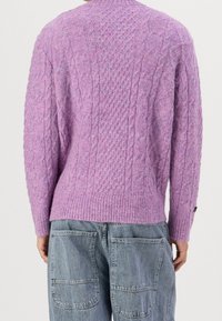 Violetti palmikkoneulepaita, jossa on resorireuna ja pitkät hihat, päällä vaaleanharmaat farkut, joissa suuret takataskut.