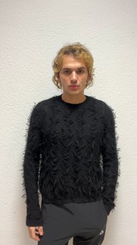 Jonge persoon met krullend blond haar draagt een zwart gestructureerd trui en donkere broek, staand tegen een effen witte muur.