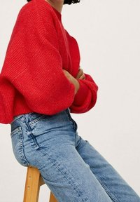 Pull en tricot rouge avec un motif texturé, associé à un jean en denim bleu clair. Le pull présente des manches surdimensionnées et une coupe décontractée.