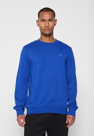 Man met kort haar draagt een effen blauwe longsleeve met een klein "4F"-logo op de borst en zwarte broek, kijkt recht vooruit.