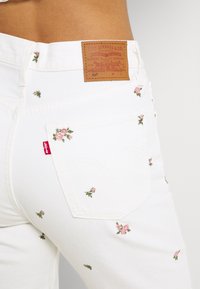 Weiße Jeans mit bestickten rosa Blumen und grünen Blättern. Verfügt über ein braunes Lederpatch am hinteren Bund und ein rotes Levi's-Tag.
