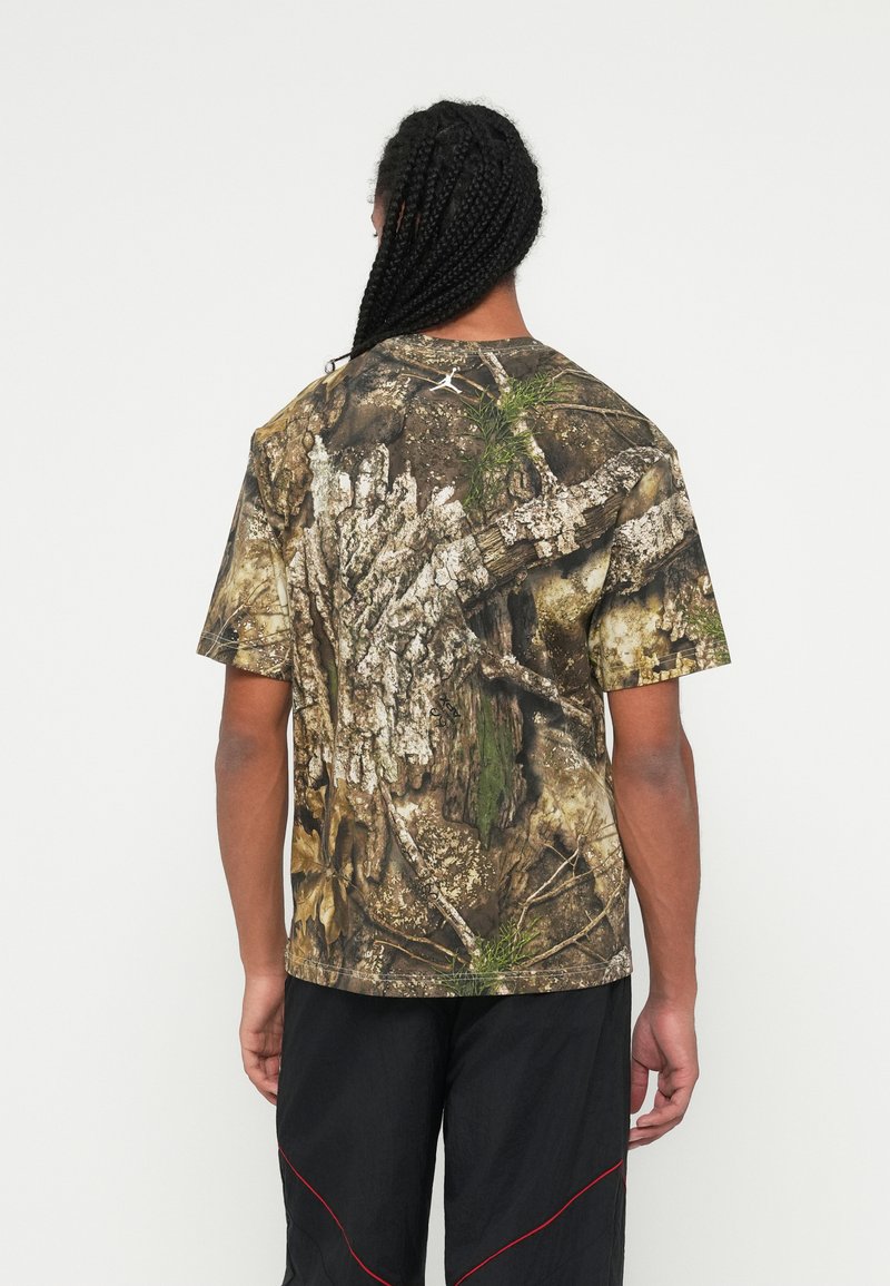 Homme avec des cheveux tressés portant un t-shirt à motif camouflage avec un design d'écorce d'arbre et un pantalon noir avec des accents rouges, de dos sur fond blanc.